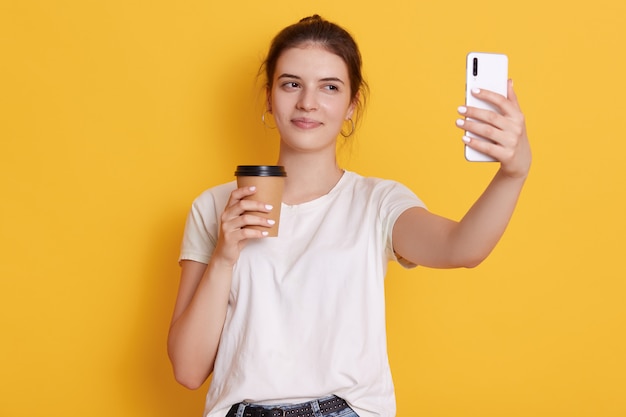 持ち帰り用のコーヒーを押しながら現代のスマートフォンを介してselfieを取る結び目を持つブルネットの若い女性