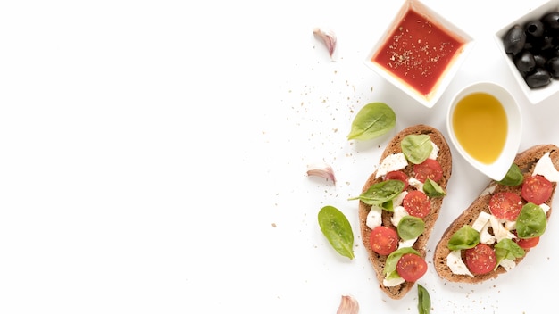 Free photo bruschetta with cheese; tomato; basil leaves topping near sauce; olives; oil and garlic clove over white surface