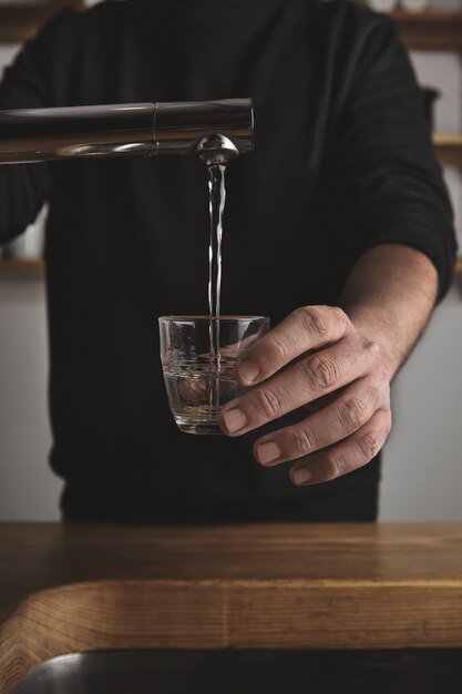 Brutal barista in black sweatshot behind thick wooden table fills small tranparent glass with water under silver metal tap in cafe shop.