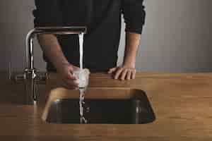 Free photo brutal barista in black sweatshot behind thick wooden table rinses small tranparent glass with water under silver metal tap in cafe shop. water drops out of glass.