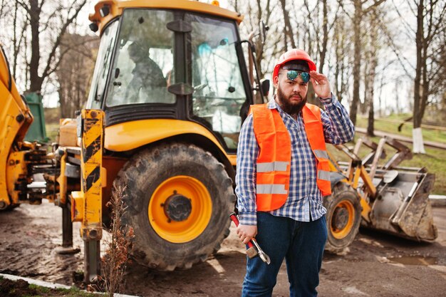 Жестокий бородатый рабочий в костюме строителя в солнцезащитных очках оранжевого шлема против трактора с разводным ключом под рукой