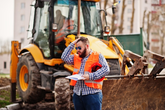 Brutal beard worker man suit construction worker in safety orange helmet sunglasses against traktor with plan paper at hands