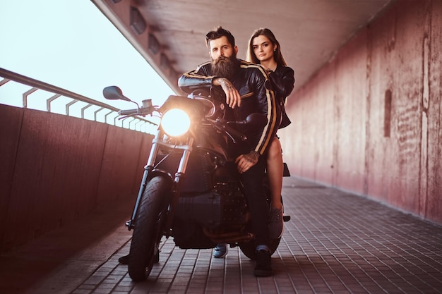 Brutal bearded biker in black leather jacket and sensual brunette girl sitting together on a custom-made retro motorcycle on a footway under a bridge.