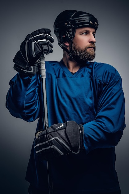 안전 헬멧을 쓴 잔인한 수염 아이스하키 선수가 회색 배경에 게임 스틱을 들고 있습니다.