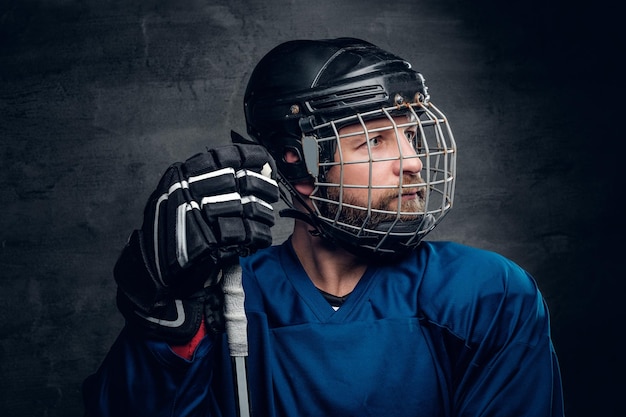 A brutal bearded ice-hockey player in safety helmet holds the gaming stick on grey vignette background.