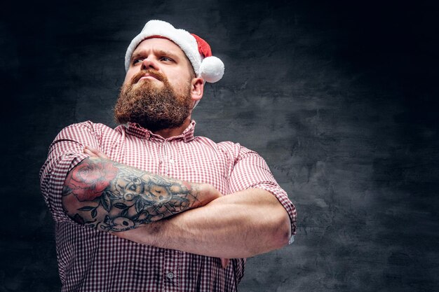 Жестокий бородатый мужчина с татуированной рукой в новогодней шапке Санты.