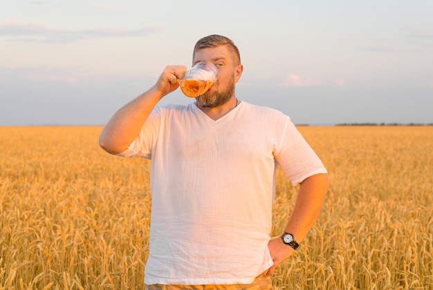 夕日の光線で麦畑に立ってビールを飲む残忍なひげを生やした男。男は泡立つ飲み物を楽しんでいます。暖かい夏の日。天然物。