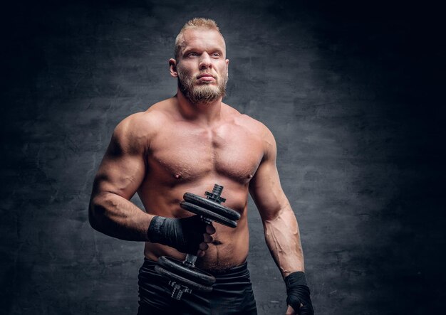 The brutal bearded muscular male holds dumbbell over grey vignette background.