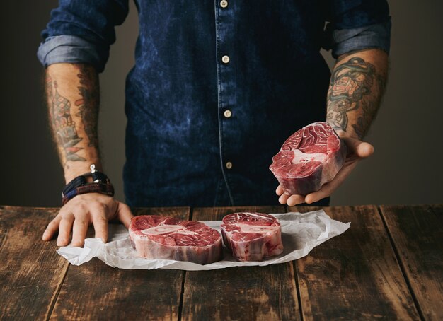 Brutal butcher with tattooed hands offers a piece of great raw meat steak on camera, other steaks in white craft paper on vintage old wooden grunge table. unrecognizable