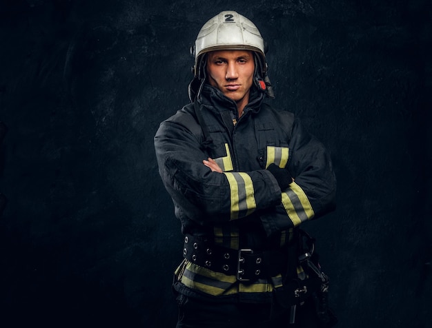 Brutal fireman in uniform posing for the camera standing with crossed arms and confident look. Studio photo against a dark textured wall