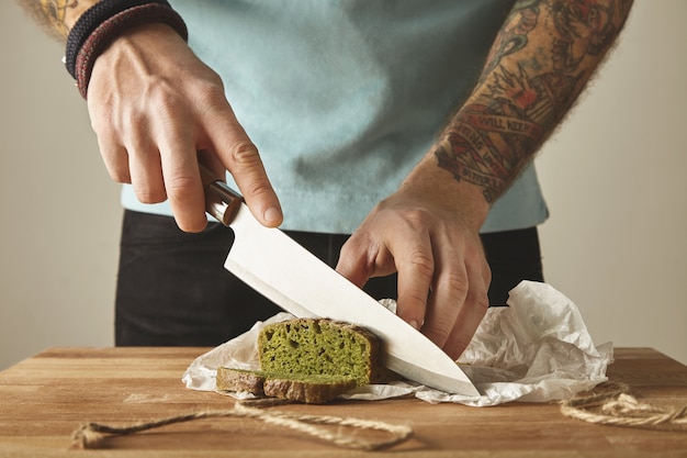 残忍な男の入れ墨の手は、スライスにヴィンテージナイフで健康的なほうれん草自家製の緑の素朴なパンをカットします。木の板の白いテーブル