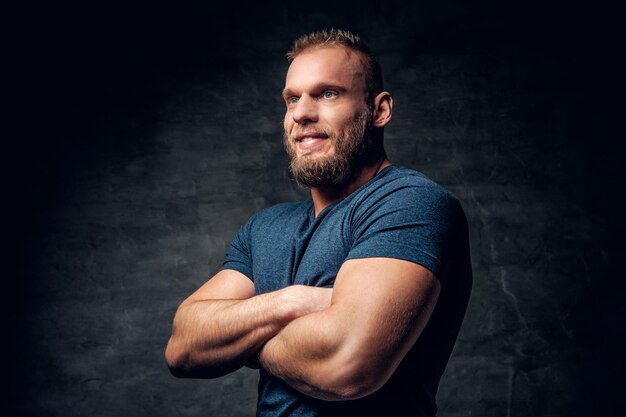 Brutal muscular male with crossed arms, dressed in a blue t shirt over grey background.