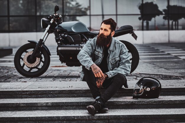 데님 재킷을 입은 잔인한 생각에 잠긴 남자가 유리 건물 근처에 있는 그의 오토바이 근처 계단에 앉아 있습니다.