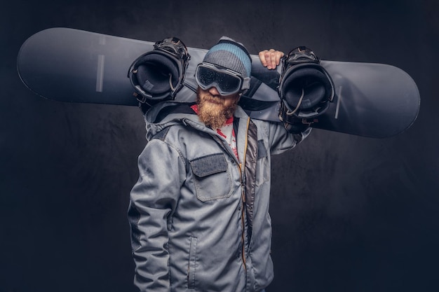 Brutal redhead snowboarder with a full beard in a winter hat and protective glasses dressed in a snowboarding coat holds a snowboard on his shoulder in a studio. Isolated on gray background.