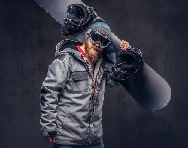 Brutal redhead snowboarder with a full beard in a winter hat and protective glasses dressed in a snowboarding coat holds a snowboard on his shoulder in a studio. Isolated on gray background.