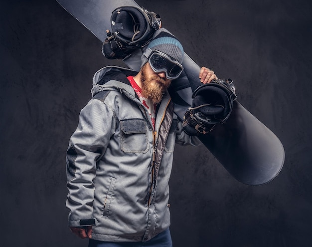 Brutal redhead snowboarder with a full beard in a winter hat and protective glasses dressed in a snowboarding coat holds a snowboard on his shoulder in a studio. Isolated on gray background.