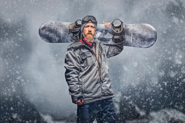 冬用帽子をかぶった完全なあごひげとスノーボードコートを着た保護メガネを身に着けた残忍な赤毛のスノーボーダーが、カメラを見て、山を背景にスノーボードでポーズをとっています。