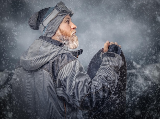 Foto gratuita uno snowboarder rosso brutale con la barba piena in un cappello invernale e occhiali protettivi vestito con un cappotto da snowboard in posa con lo snowboard sullo sfondo delle montagne.
