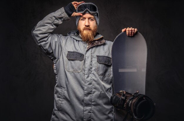 Жестокий рыжий сноубордист с полной бородой в зимней шапке и защитных очках, одетый в пальто для сноуборда, позирует со сноубордом в студии. Изолированные на сером фоне.