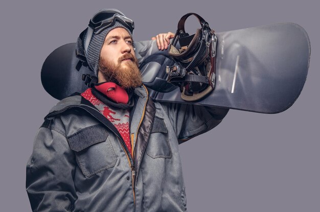 Brutal redhead snowboarder with a full beard in a winter hat and protective glasses dressed in a snowboarding coat posing with snowboard at a studio, looking away. Isolated on a gray background.