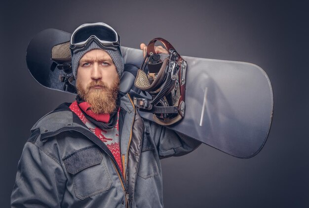 Жестокий рыжий сноубордист с полной бородой в зимней шапке и защитных очках, одетый в сноубордическую куртку, позирует со сноубордом в студии, смотрит в камеру. Изолированный на сером му