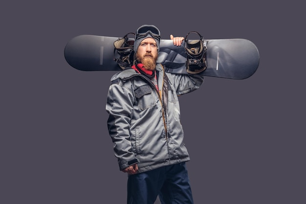 Snowboarder rosso brutale con una barba piena in un cappello invernale e occhiali protettivi vestito con un cappotto da snowboard in posa con uno snowboard in uno studio, guardando una telecamera. isolato su uno sfondo grigio