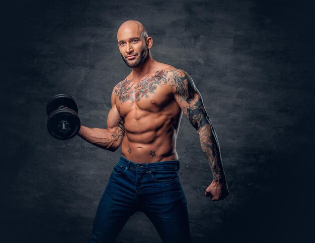 Brutal shirtless shaved head, muscular male with tattoos on his chest and arms holds dumbbell.