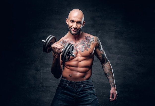 Brutal shirtless shaved head, muscular male with tattoos on his chest and arms holds dumbbell.