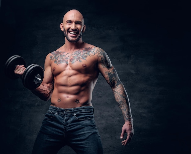 Brutal shirtless shaved head, muscular male with tattoos on his chest and arms holds dumbbell.