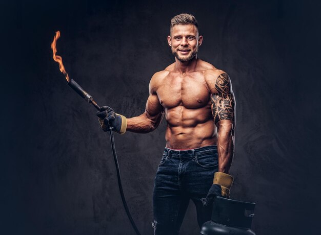 Brutal tattoed male welder with a stylish haircut and beard, with muscular body, dressed in only jeans, holds propane tank and a burning burner, standing in a studio, looking at a camera.  Isolated on