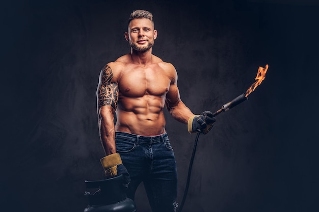 Free photo brutal tattoed male welder with a stylish haircut and beard, with muscular body, dressed in only jeans, holds propane tank and a burning burner, standing in a studio, looking with a smirk. isolated on