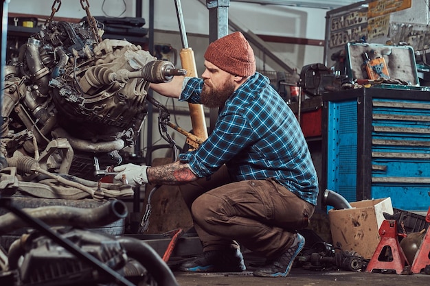 Brutal tattooed bearded mechanic specialist repairs the car engine which is raised on the hydraulic lift in the garage. Service station.