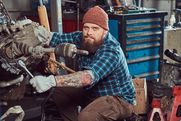 Брутальный татуированный бородатый механик ремонтирует двигатель автомобиля, который поднят на гидравлическом подъемнике в гараже. Станция обслуживания.