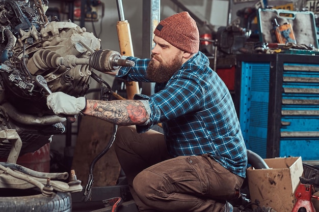Брутальный татуированный бородатый механик ремонтирует двигатель автомобиля, который поднимается на гидравлическом подъемнике в гараже. Станция обслуживания.