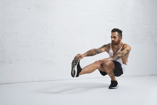 Brutal tattooed calisthenics coach shows exercise moves one leg squat, isolated on white brick wall