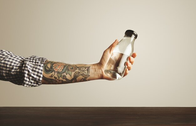 Brutal tattooed hand in folded plaid shirt holds glass transparent bottle with clean drinking water above red wooden table, isolated on white