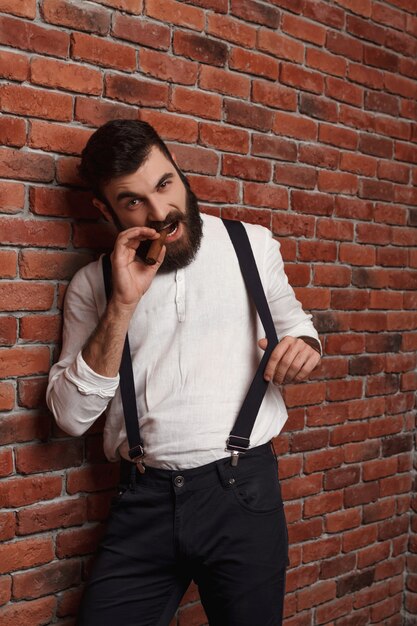 Сигары зверского молодого красивого человека куря на кирпичной стене.