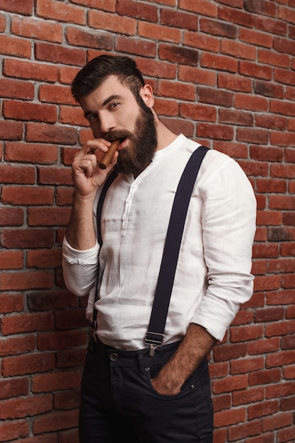 Free photo brutal young handsome man smoking cigar on brick wall.