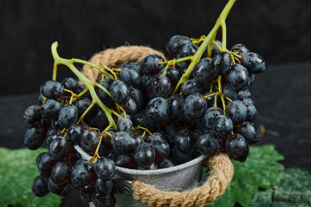 A bucket of black grapes with leaves on black surface, close up