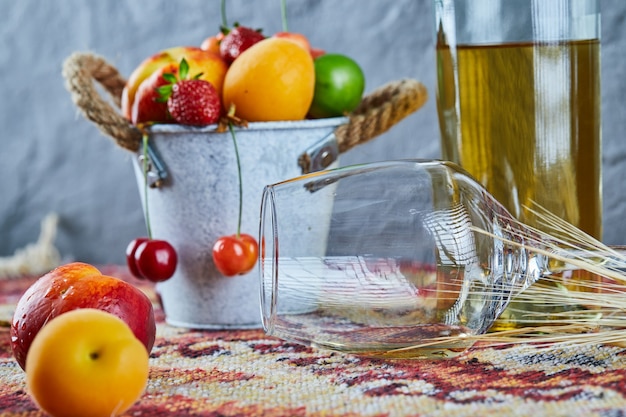 Bucket of fresh summer fruits, bottle of white wine and empty glass on carved rug.