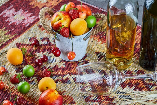 Bucket of fresh summer fruits, bottle of white wine and empty glass on carved rug.