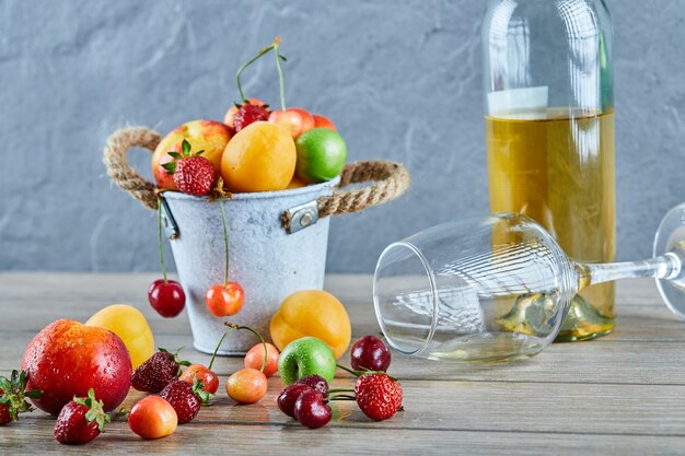 Bucket of fresh summer fruits, bottle of white wine and empty glass on wooden table.