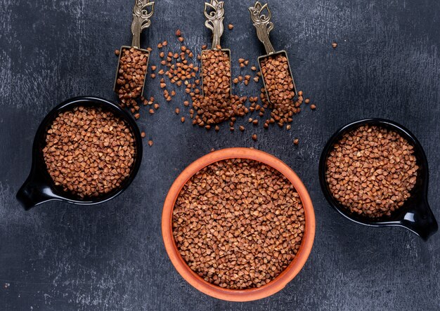 Buckwheat in a bowl and black cups and shovels top view on a dark 