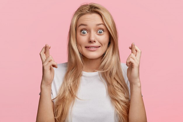 Bugged eyed blonde female model wishes greatly fortune before exam, keeps fingers crossed, dressed casually, poses against pink wall. People, facial expressions, emotions and great desire