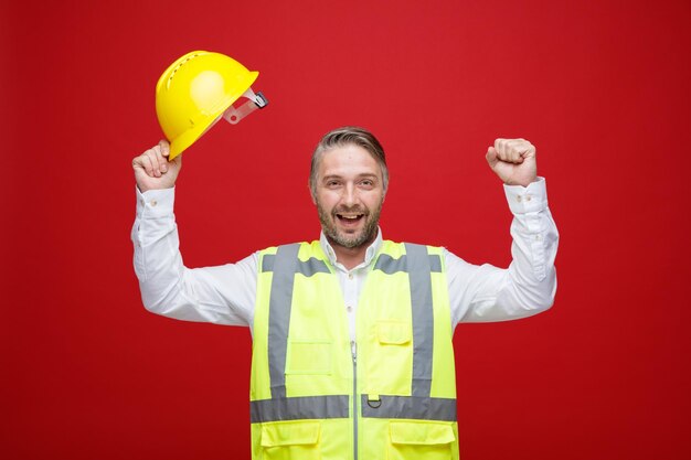 Builder man in construction uniform holding his safety helmet happy and excited raising clenched fists smiling broadly standing over red background