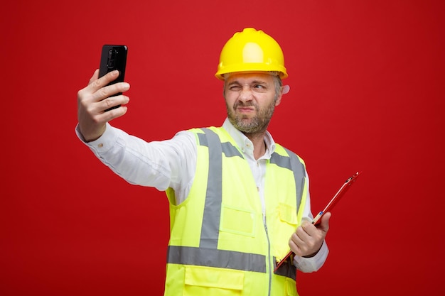 Builder man in construction uniform and safety helmet holding clipboard doing selfie using smartphone looking displeased making wry mouth standing over red background
