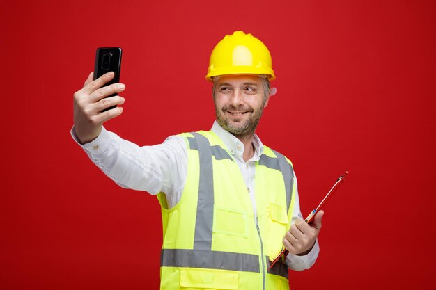 Builder man in construction uniform and safety helmet holding clipboard doing selfie using smartphone smiling cheerfully happy and pleased standing over red background