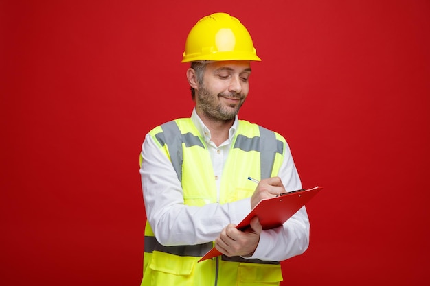 Мужчина-строитель в строительной форме и защитном шлеме, держащий буфер обмена, выглядит уверенно, делая заметки, стоя на красном фоне