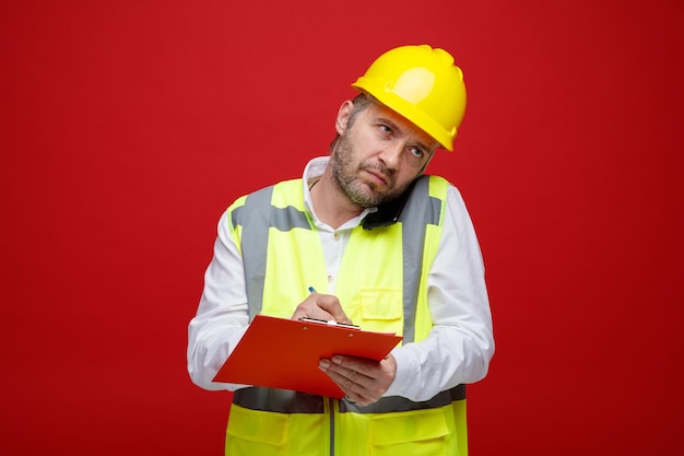 Builder man in construction uniform and safety helmet holding clipboard looking displeased while talking on mobile phone making notes standing over red background