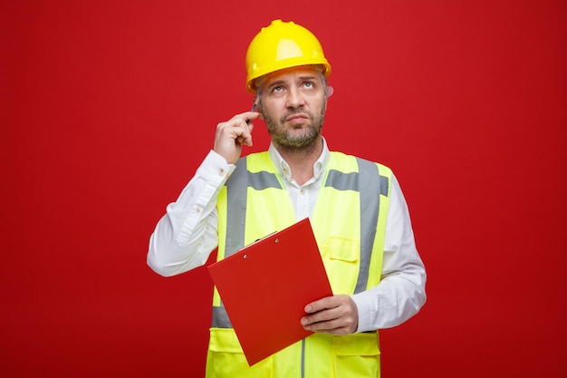 Builder man in construction uniform and safety helmet holding clipboard looking up puzzled scratching his head standing over red background